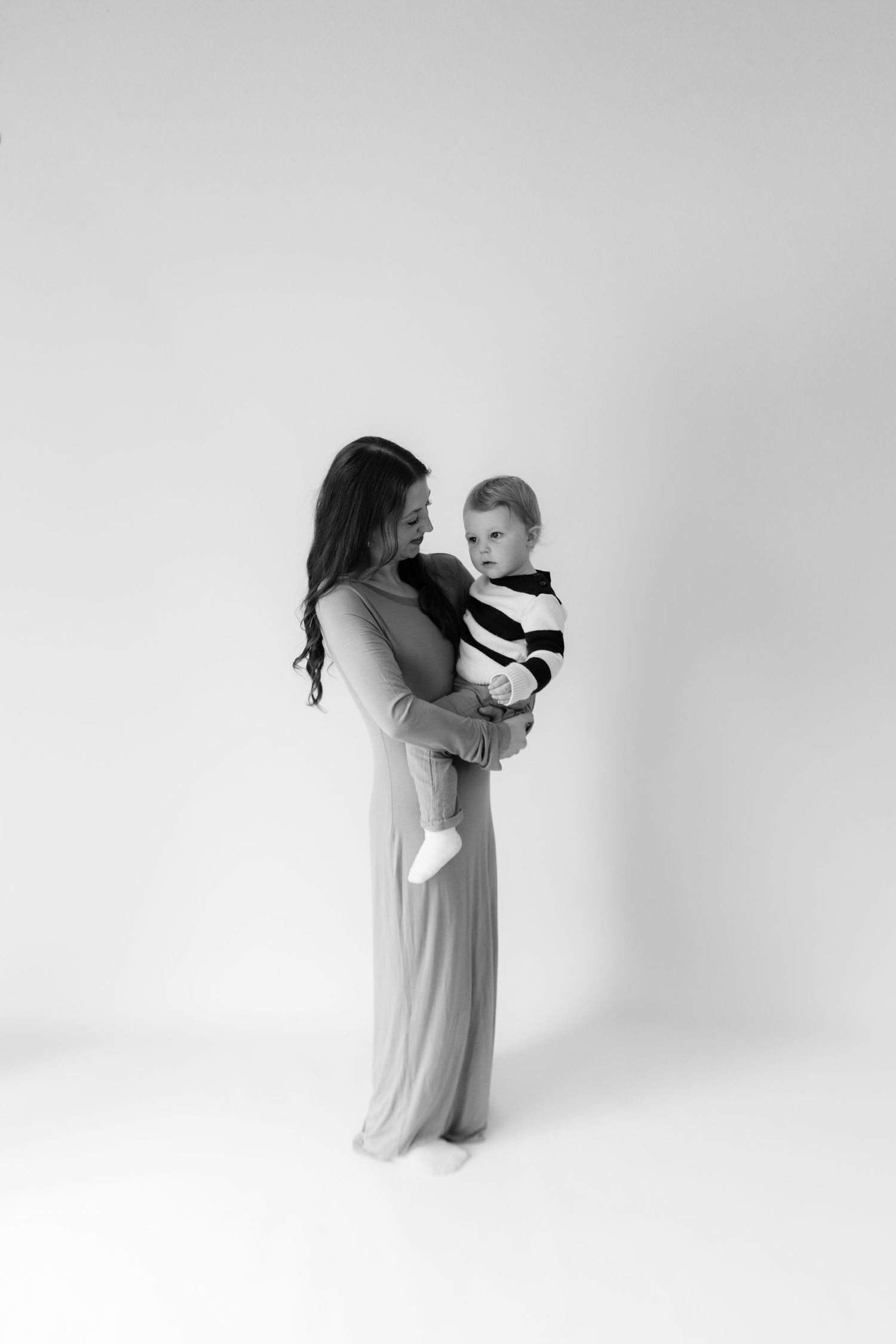 Black and white photo of a mother in a long dress holding her baby boy in a striped outfit in a studio.