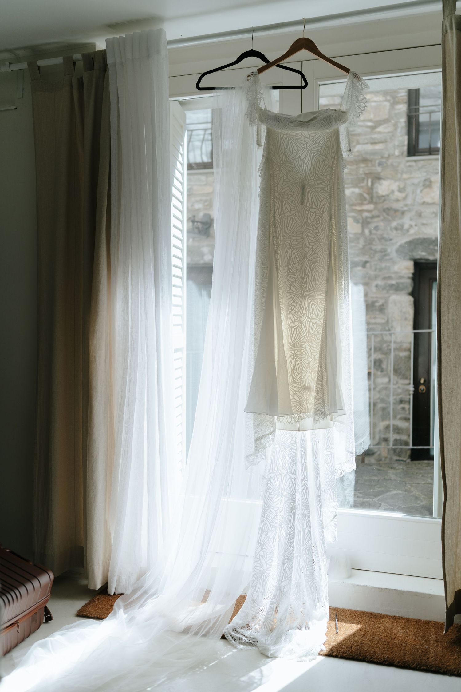 White lace wedding dress hangs in window light against stone wall creating ethereal bridal preparation scene.