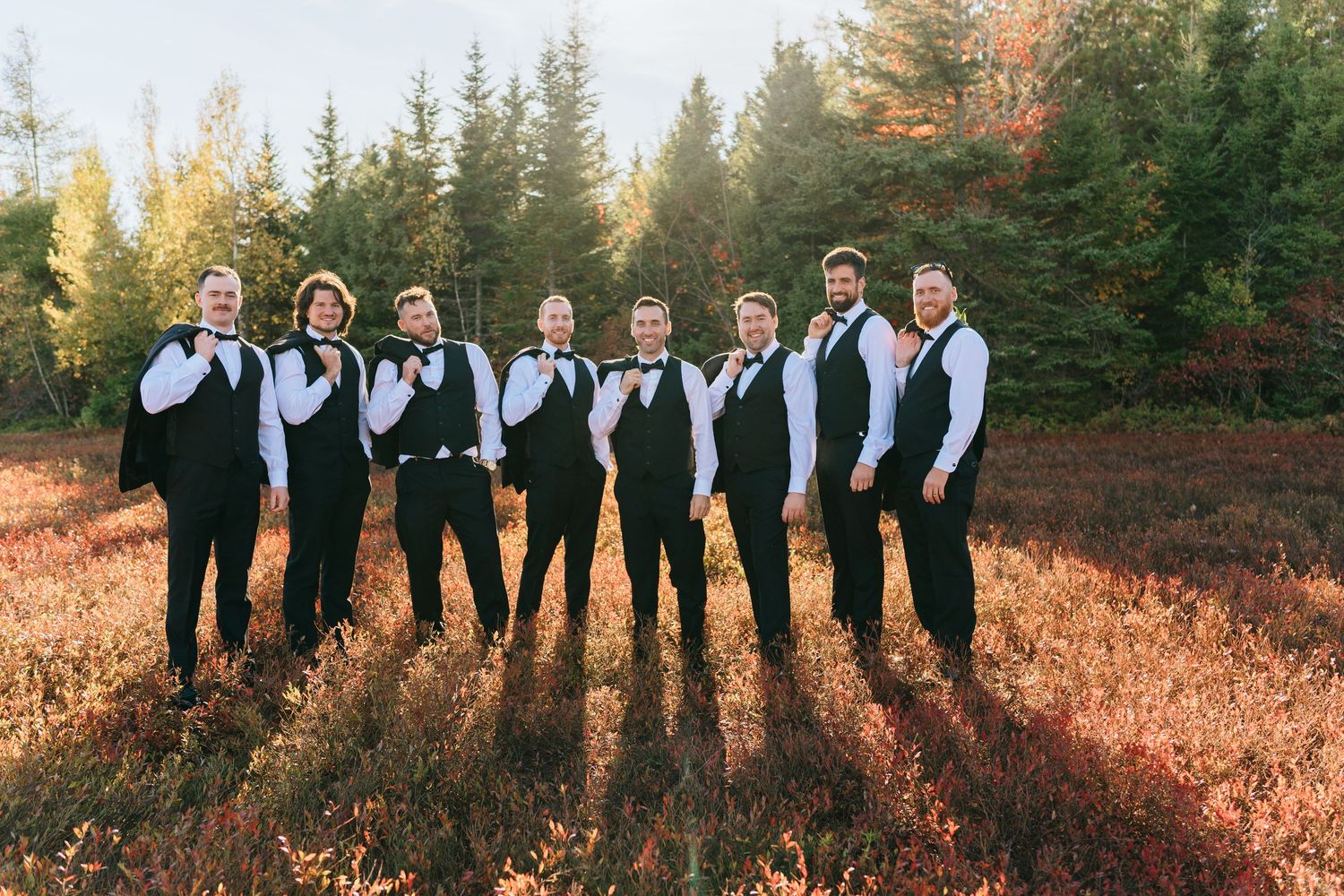 Groomsmen in black vests and bow ties posing in a colorful autumn forest clearing.