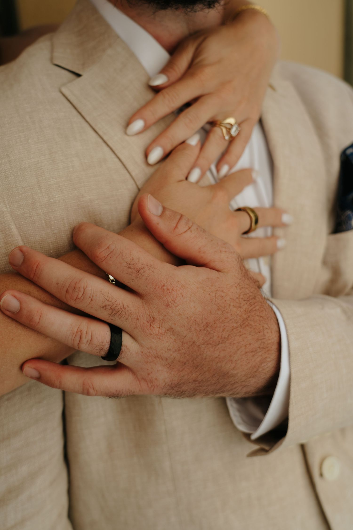 Close-up of hands wearing wedding rings against a light beige suit jacket in Italian village