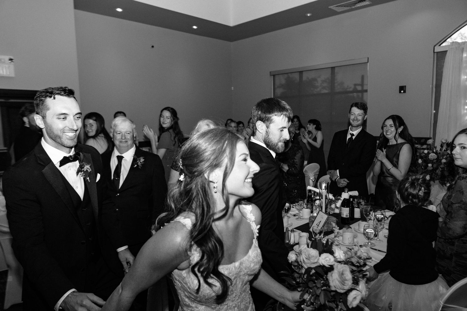 Black and white wedding reception photo of bride and groom smiling as guests celebrate around them.