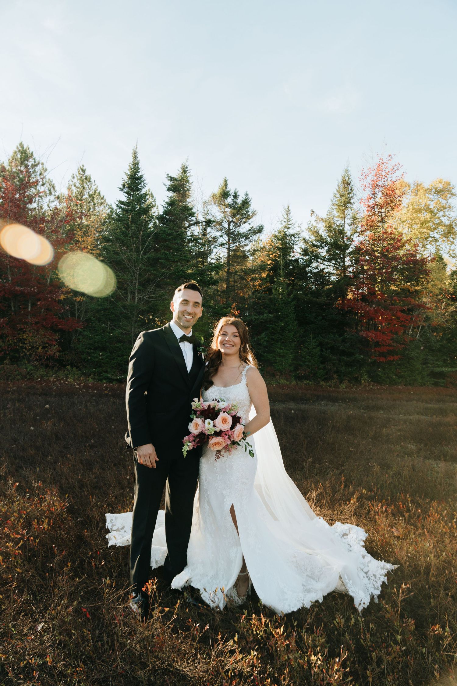 Bride and groom posing in autumn field with colorful fall foliage and pine trees backdrop on their wedding day.