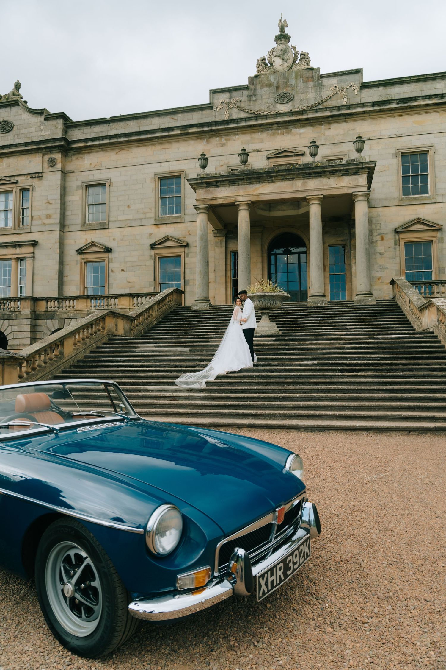 Bride and groom embrace on grand estate steps beside a classic blue MG convertible on their wedding day.
