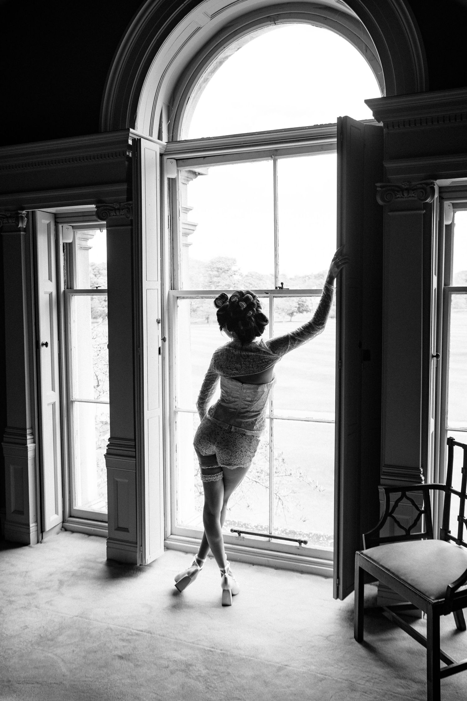 Black and white photo of a woman in lingerie and heels standing at an elegant arched window in a grand interior.