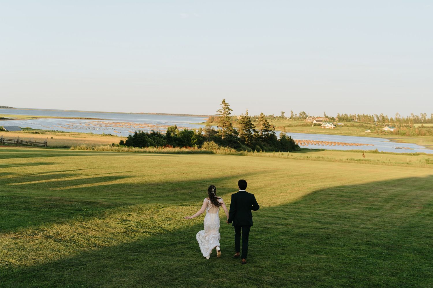 A couple in wedding attire walks across a scenic grassy field during golden hour.