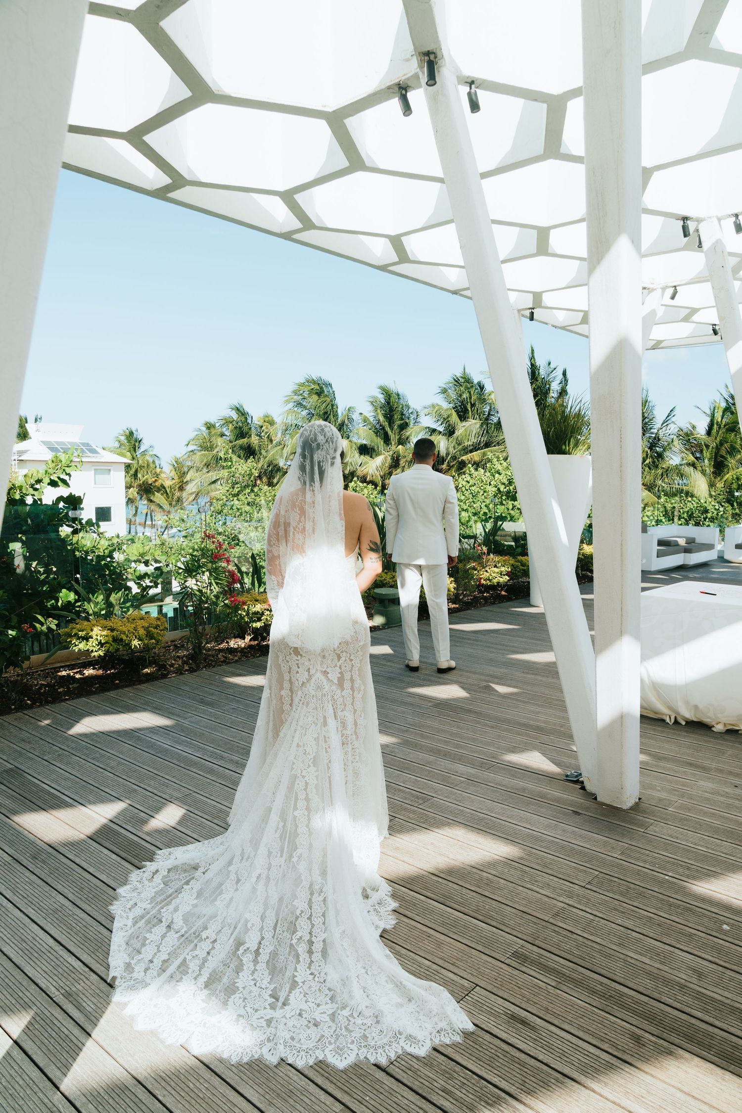 Bride in lace wedding gown and veil stands on wooden deck under white geometric pergola, groom walks ahead near tropical palms.