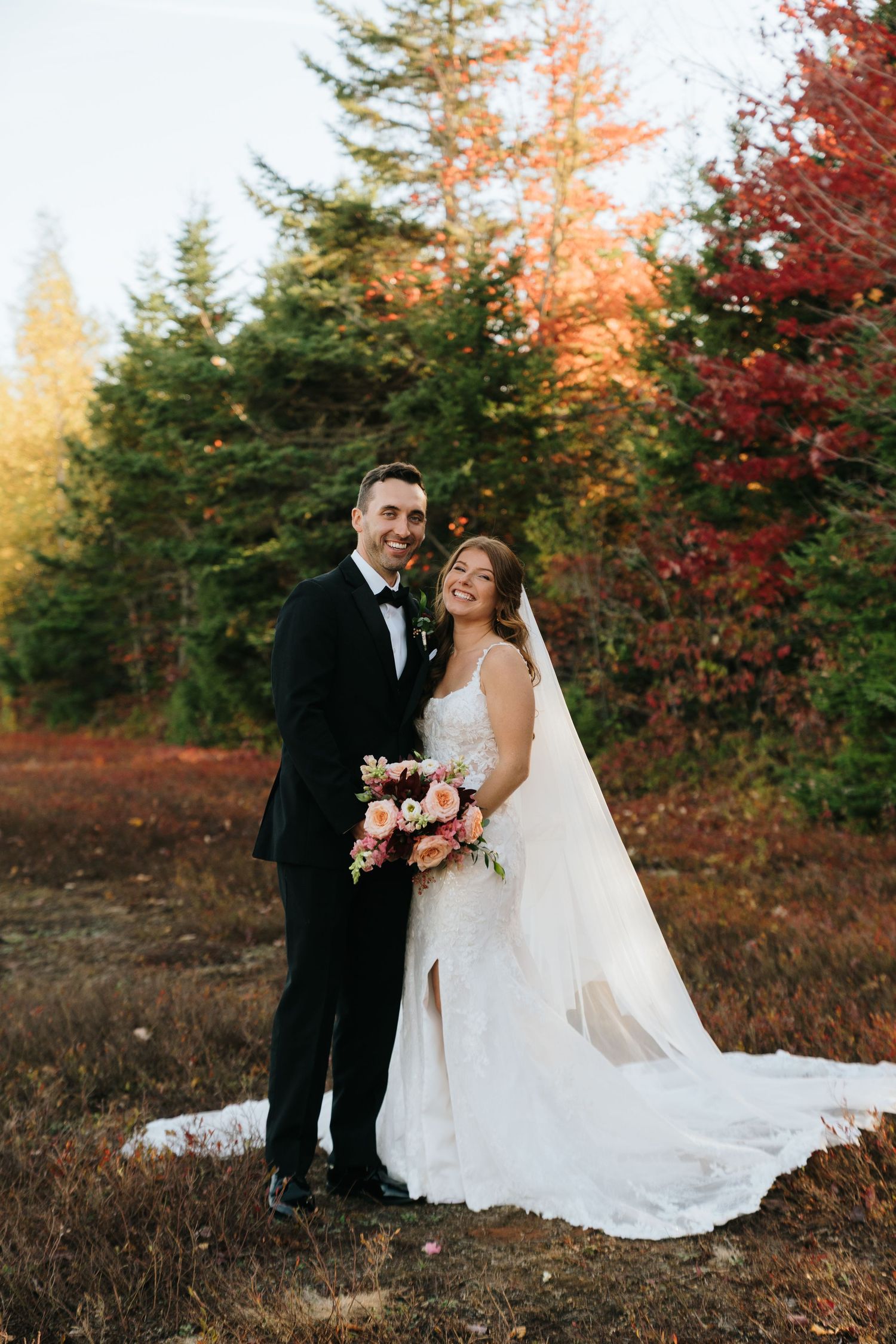 A couple shares a romantic moment in their wedding attire against a backdrop of autumn foliage and evergreen trees.