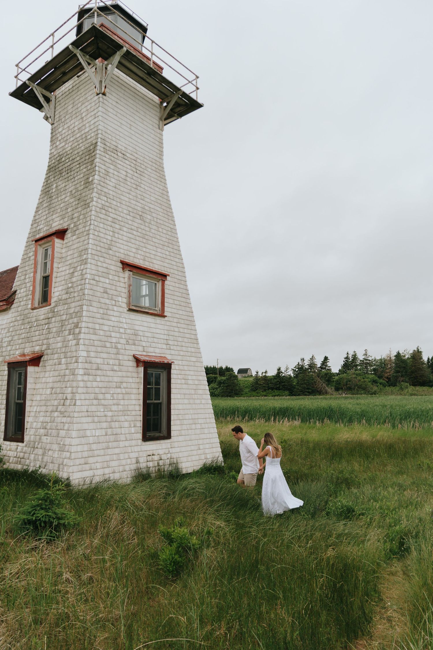 A romantic couple poses near a white lighthouse in a grassy field on an overcast day.
