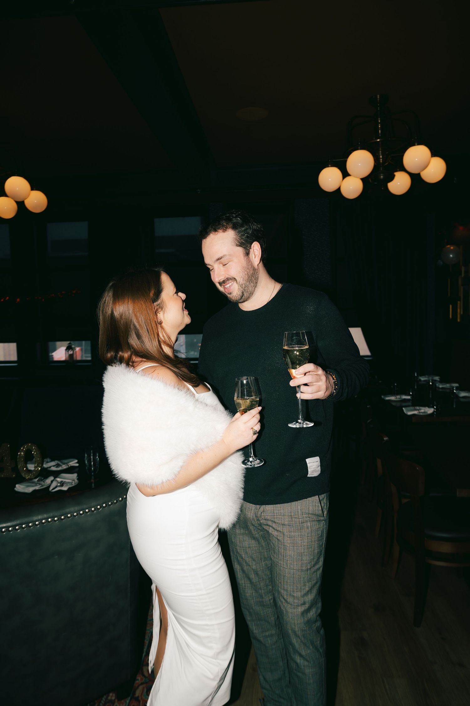 Couple sharing a romantic toast with white wine glasses at an elegant dark restaurant, woman in white dress with fur wrap.