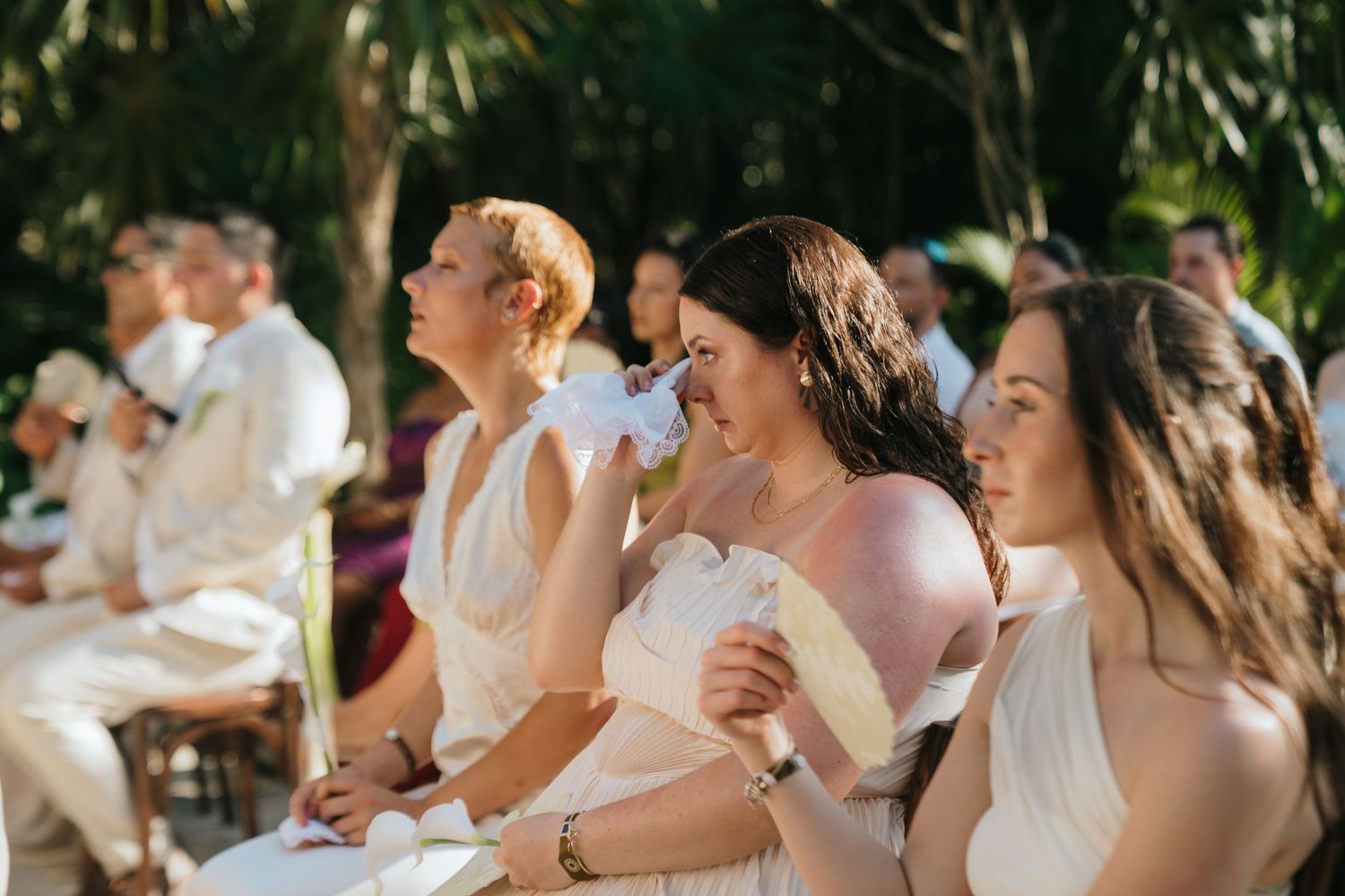 Bridesmaids in white dresses fan themselves during an outdoor wedding ceremony surrounded by lush tropical greenery.