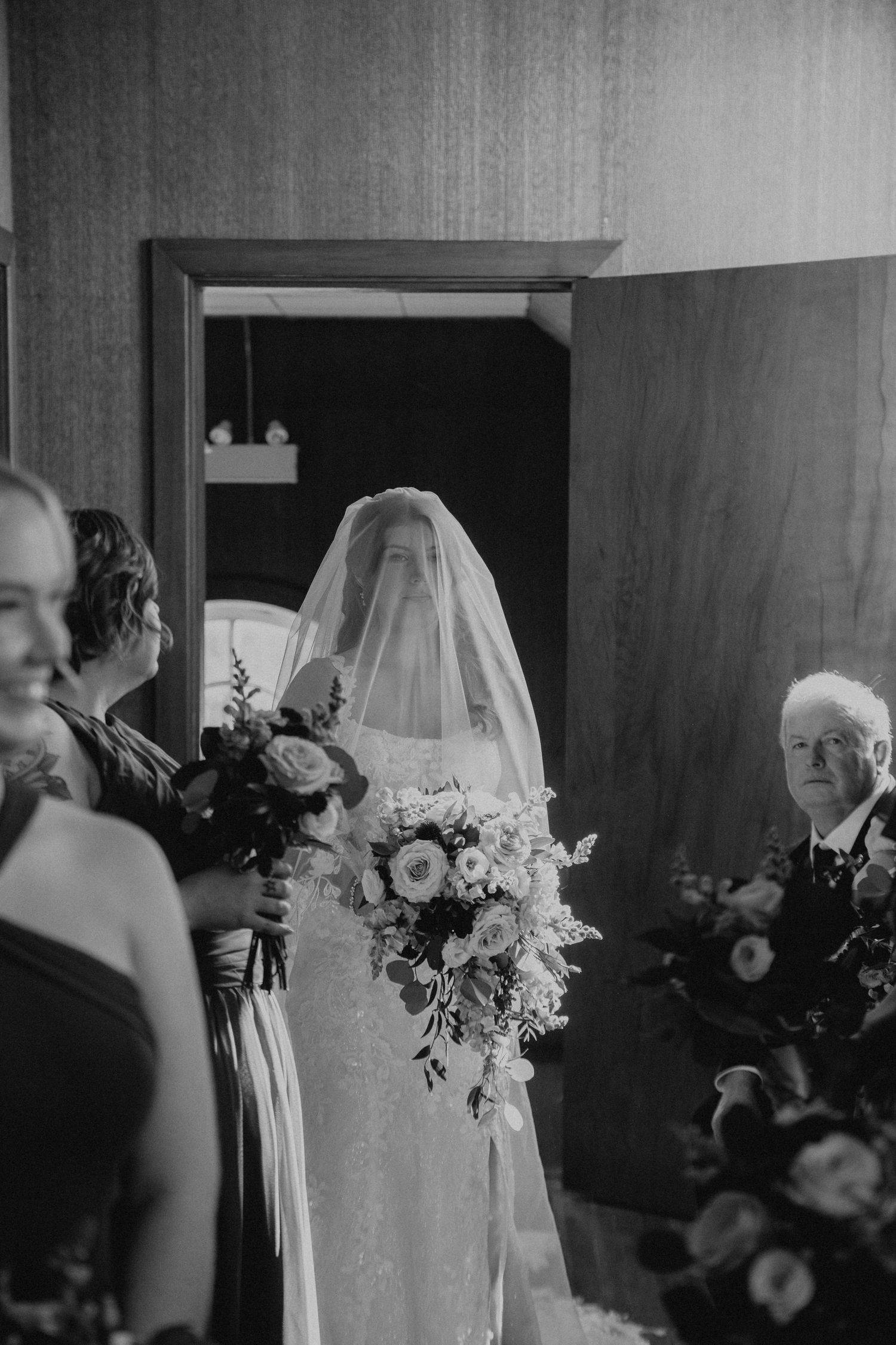 Black and white photo of a veiled bride holding a bouquet walking down the aisle with her father at a wedding.