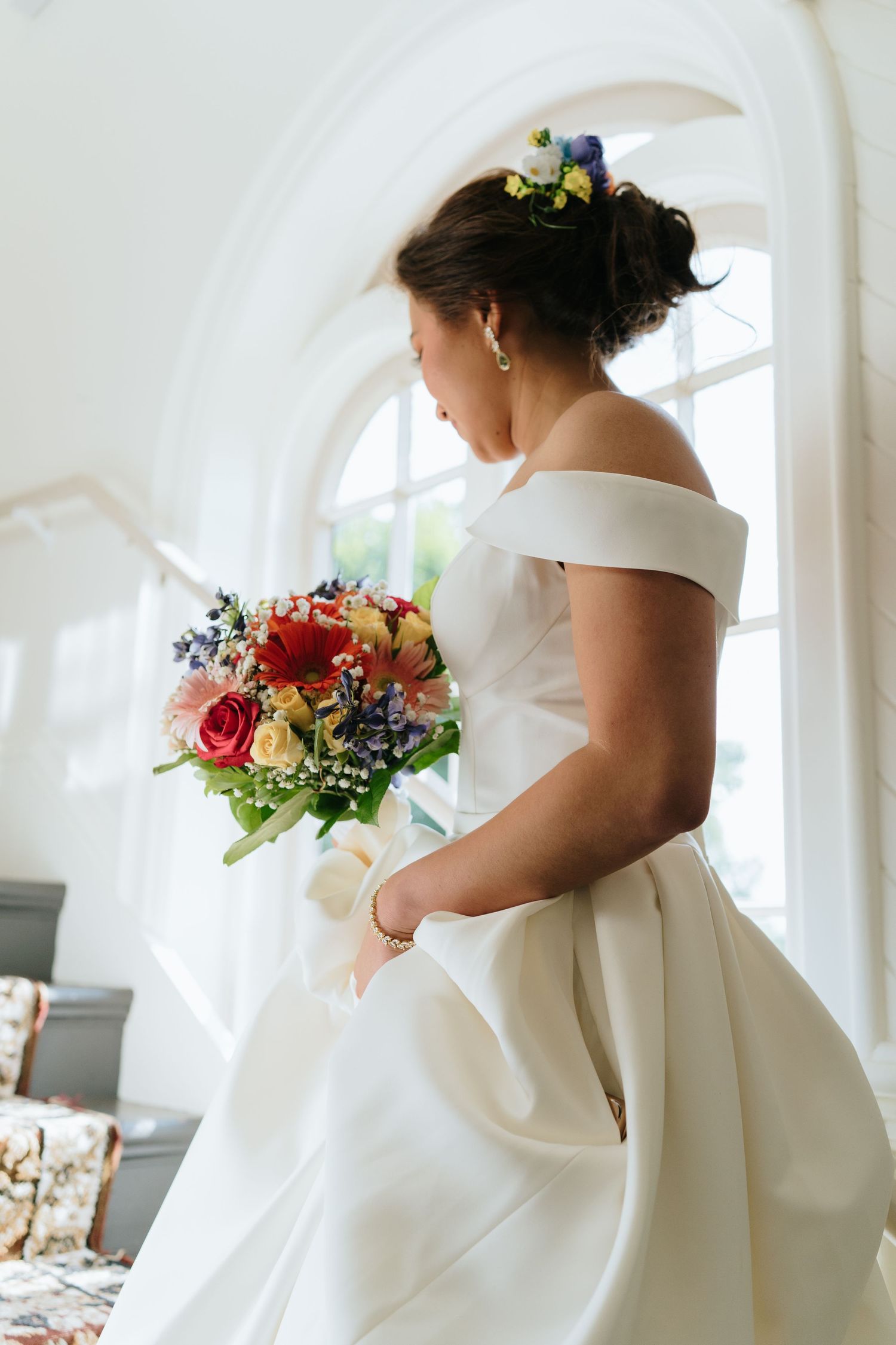A wedding dress with an elegant off-shoulder neckline and colorful floral bouquet in natural light.