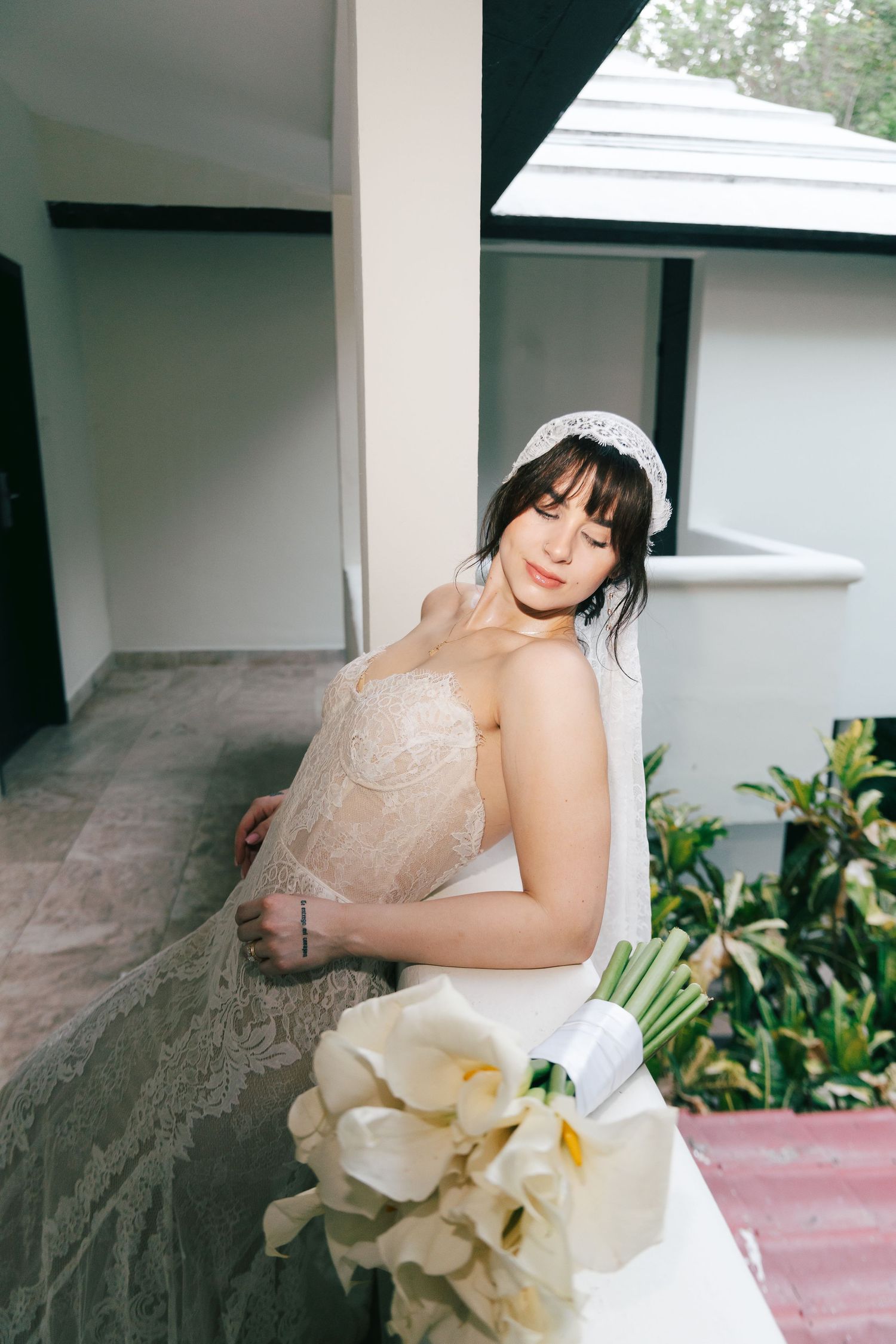 Elegant bride in lace wedding dress with veil headpiece holding white calla lily bouquet on outdoor balcony.
