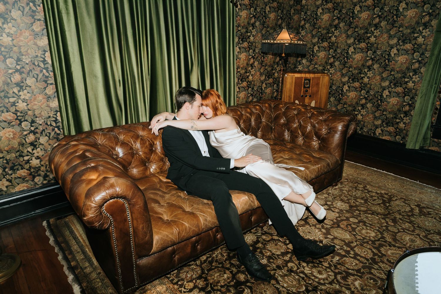 Couple resting together on a vintage leather chesterfield couch in an ornate room.