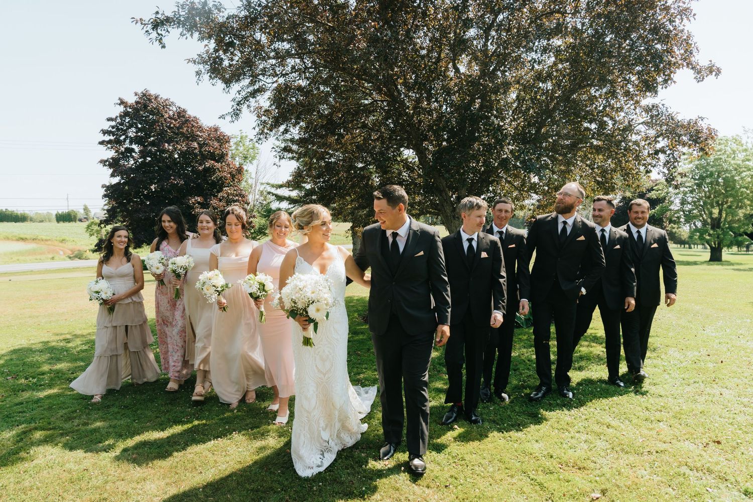 Wedding party in formal attire poses under trees on a sunny summer day.