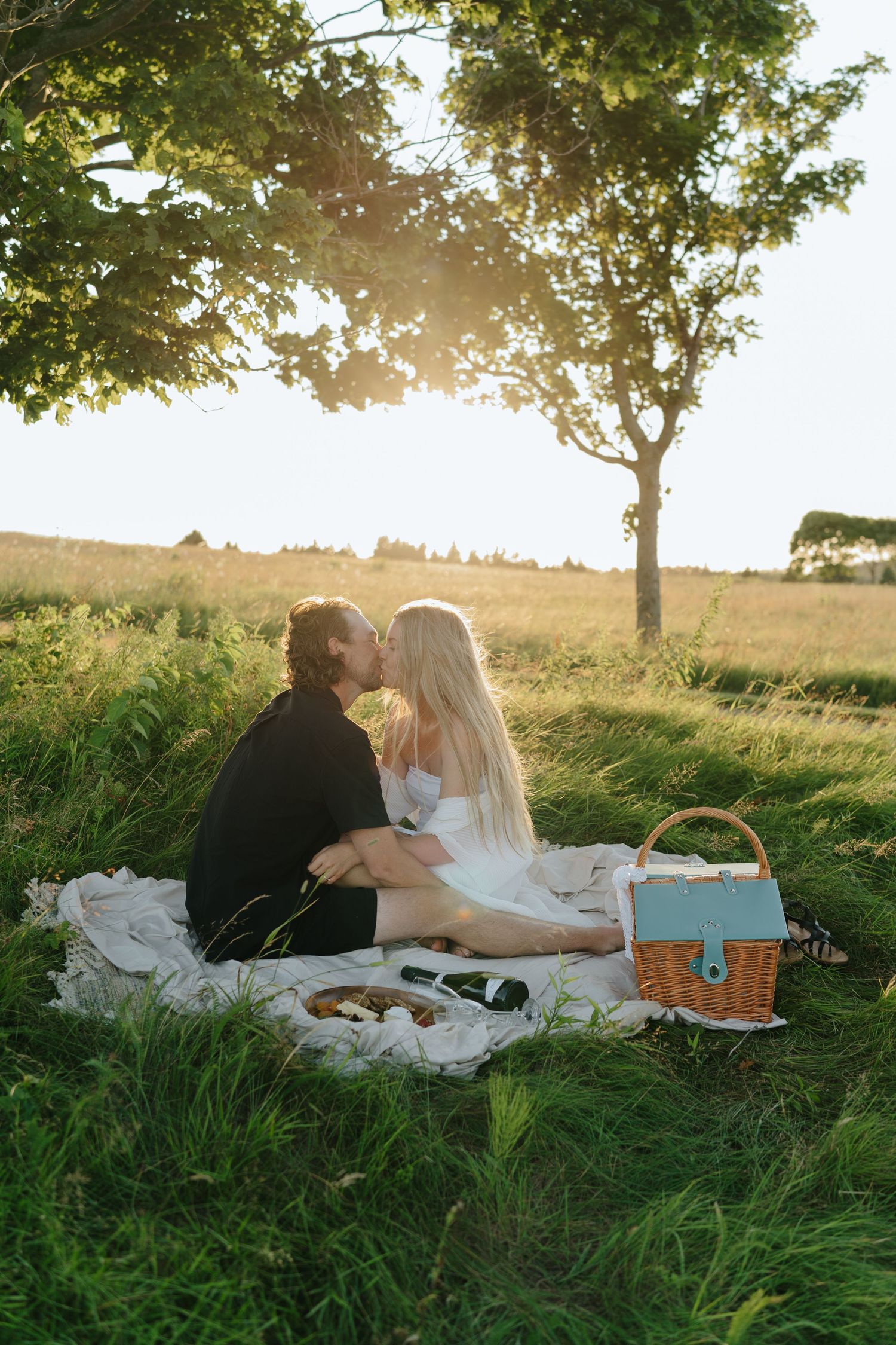 A couple enjoys a romantic sunset picnic on a blanket in a grassy hilltop meadow during golden hour.