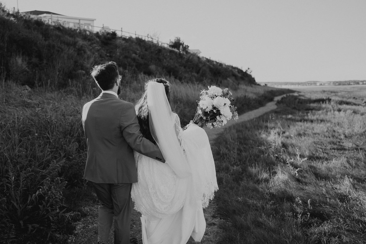 Black and white wedding photograph capturing a couple walking along a scenic hillside path.