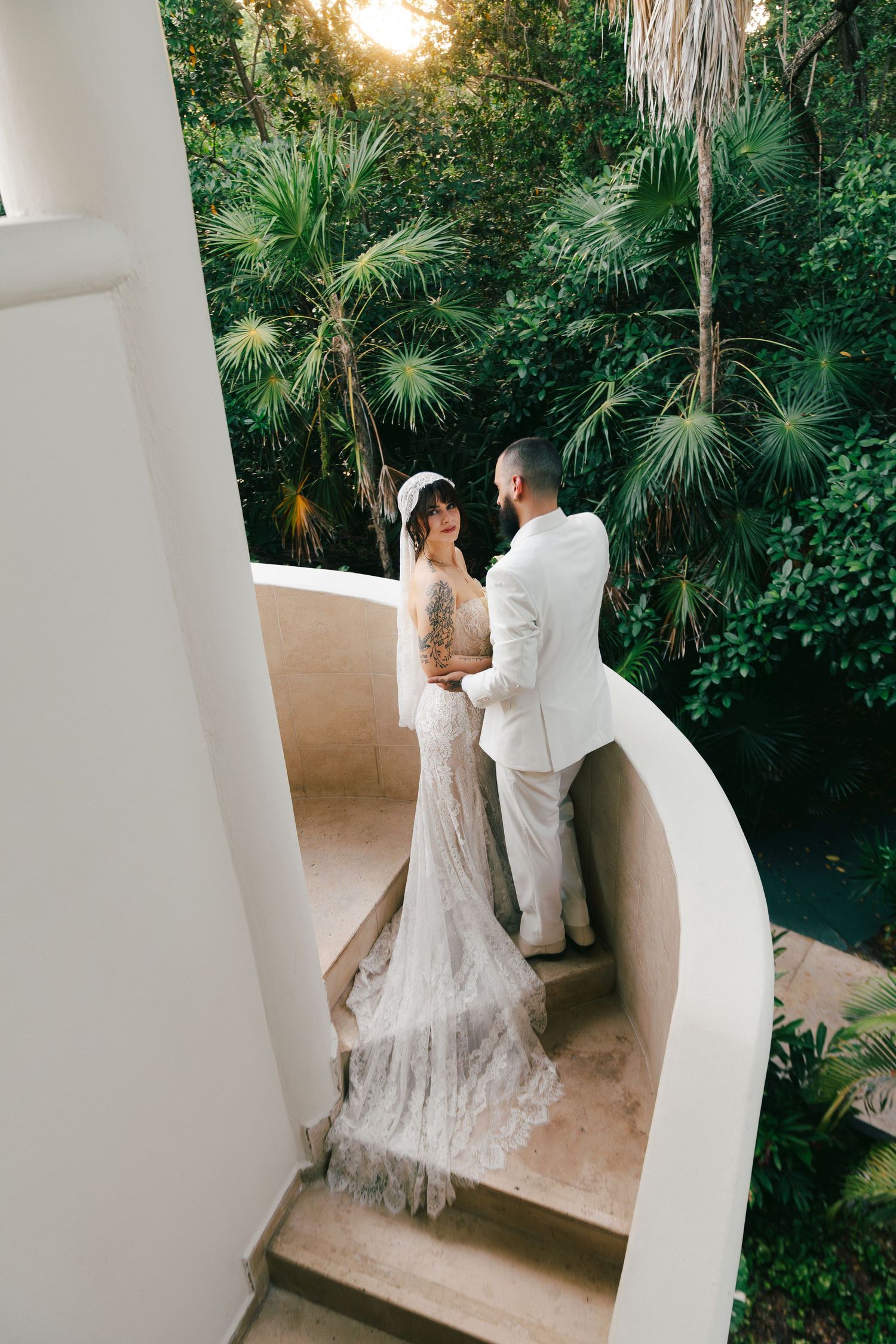 Bride in lace gown and groom in white suit sharing romantic moment on curved white staircase balcony with tropical greenery.