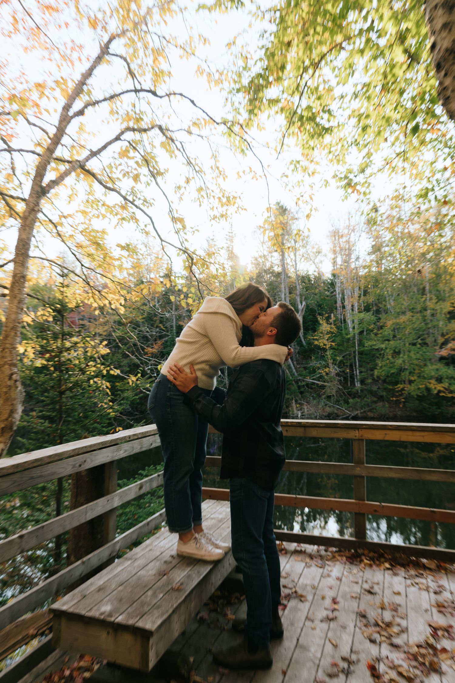 A couple shares an intimate moment on a wooden deck overlooking autumn trees.
