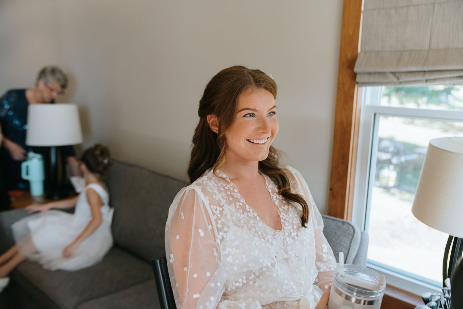 Smiling bride in lace wedding dress getting ready with bridesmaids in background near window.