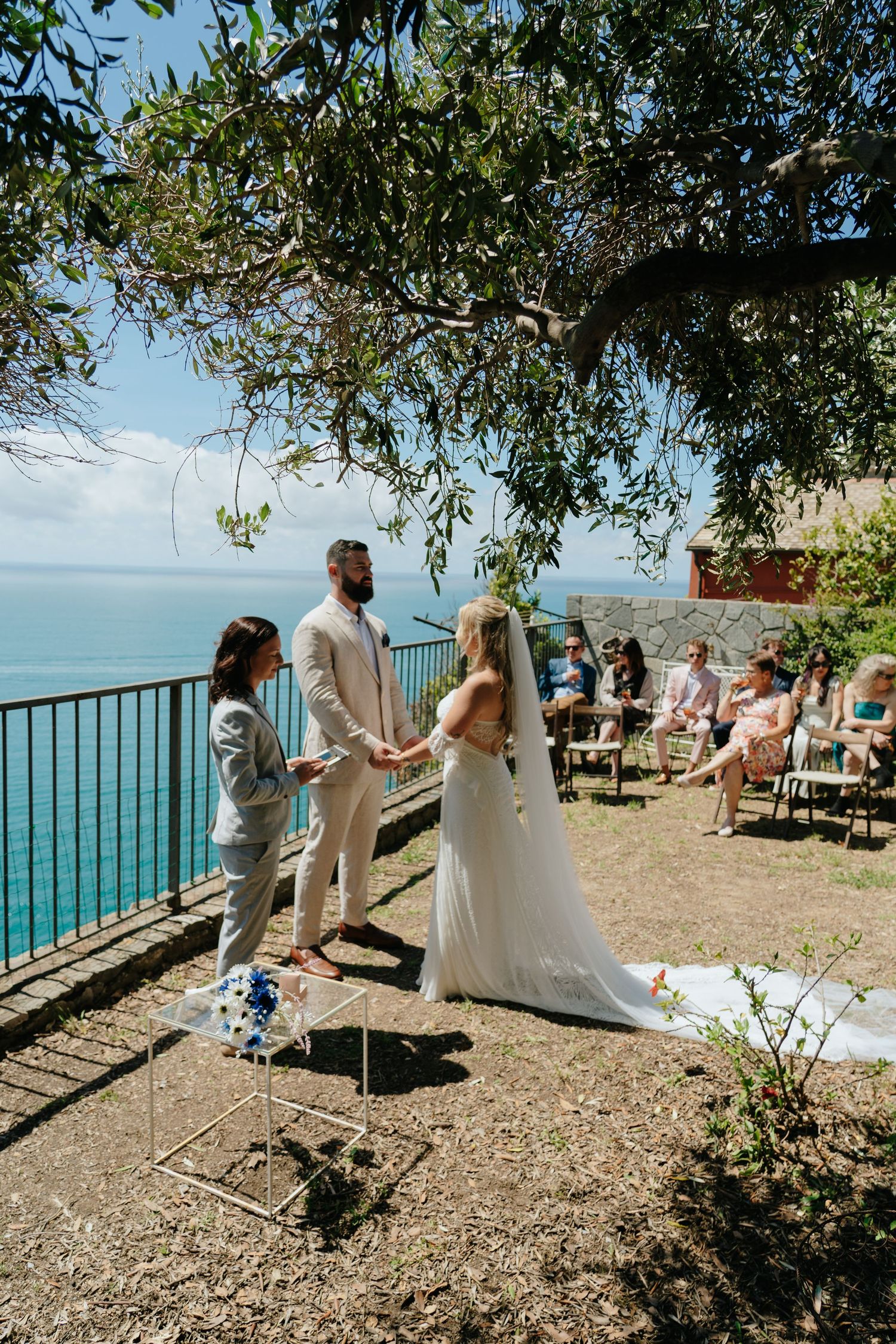 Wedding ceremony guests gather on a scenic coastal overlook with turquoise waters and blue skies in the background.