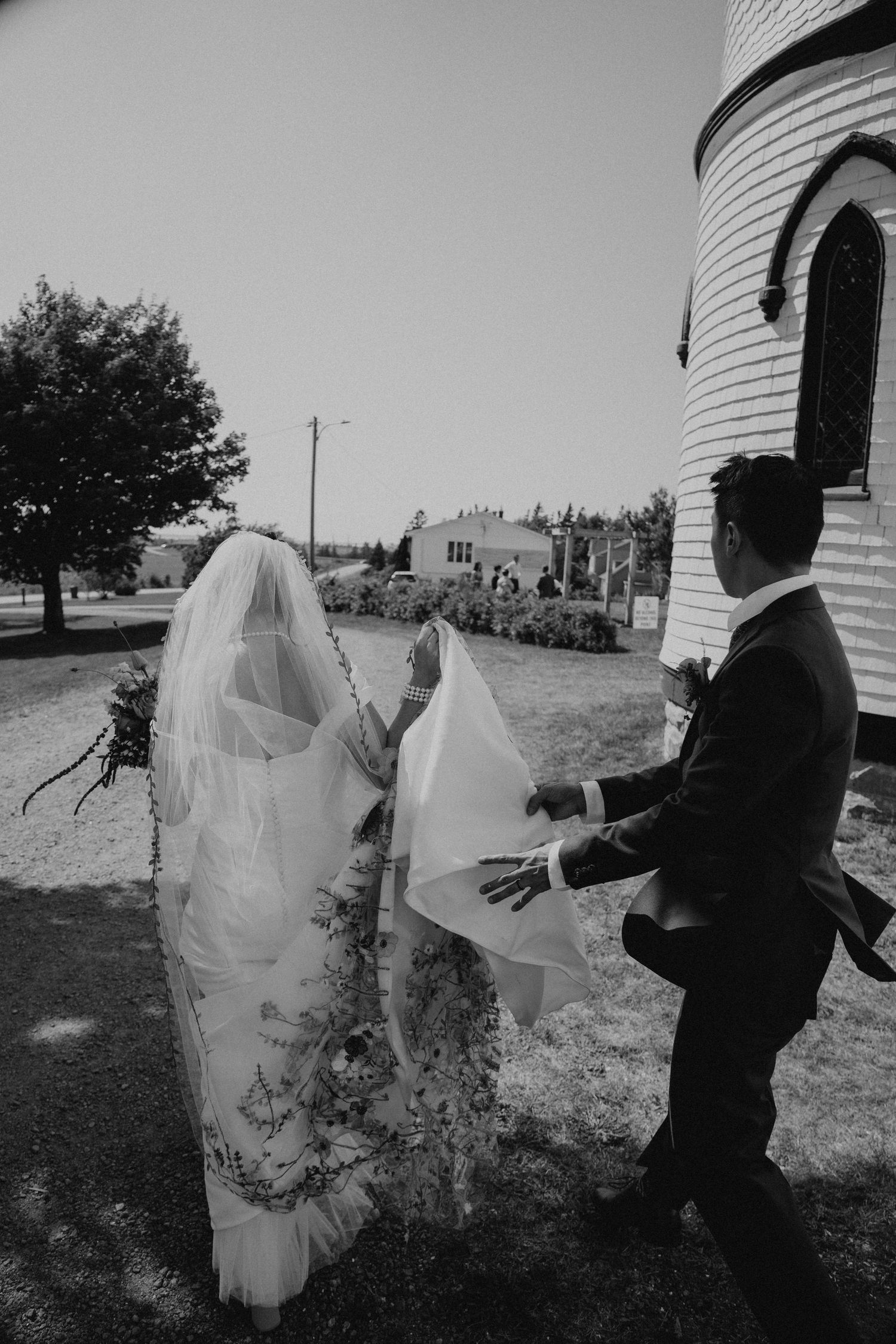 Black and white photo of a couple's playful moment with a flowing veil near a lighthouse.