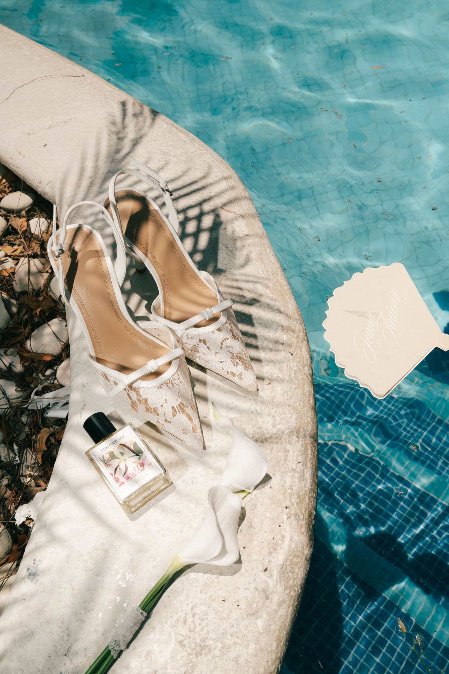 White bridal heels and perfume bottle displayed on stone edge beside turquoise pool with envelope, wedding flat lay.