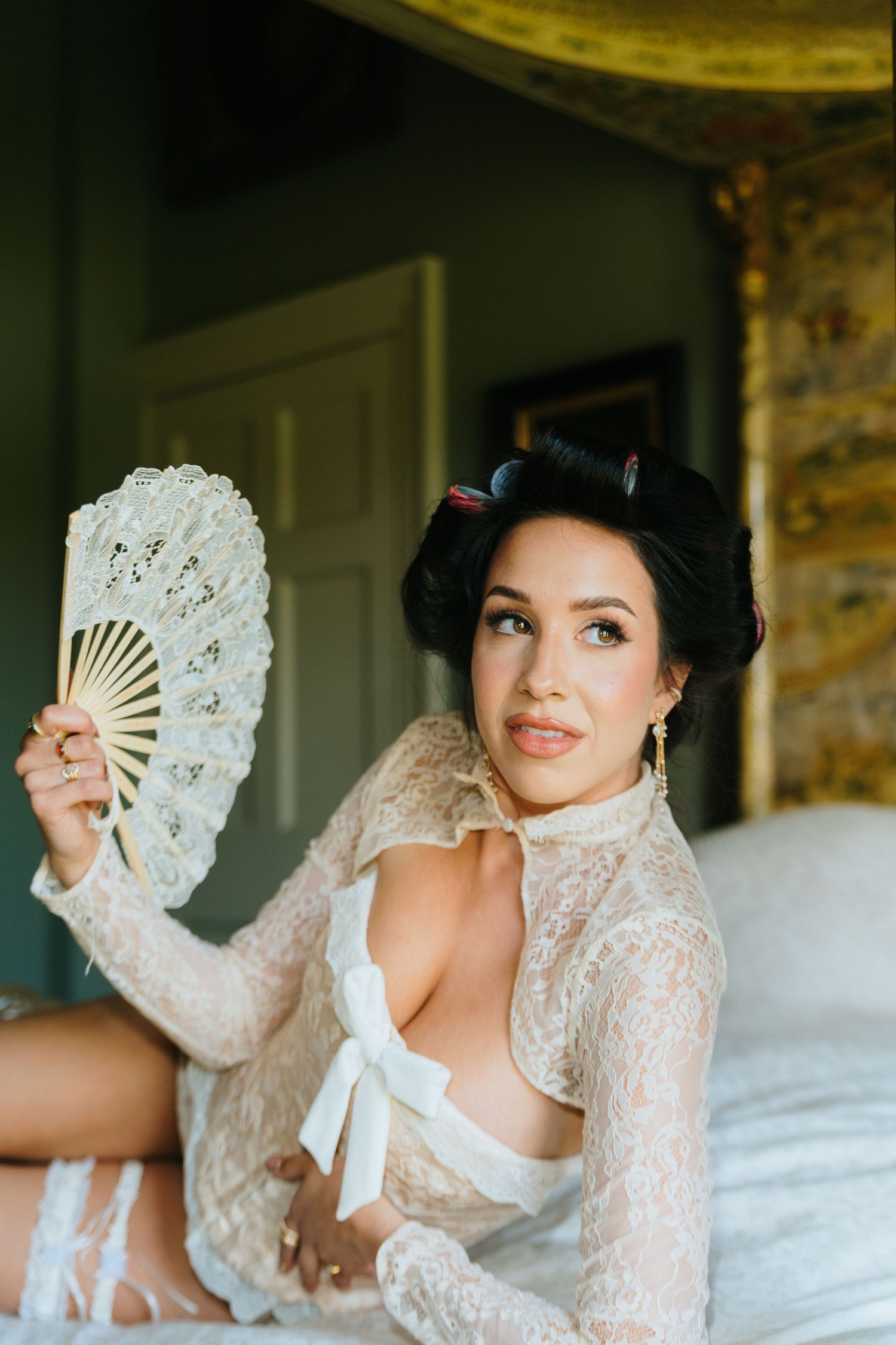 Elegant bride in lace robe holding a decorative fan, sitting on bed with vintage-inspired updo hairstyle.