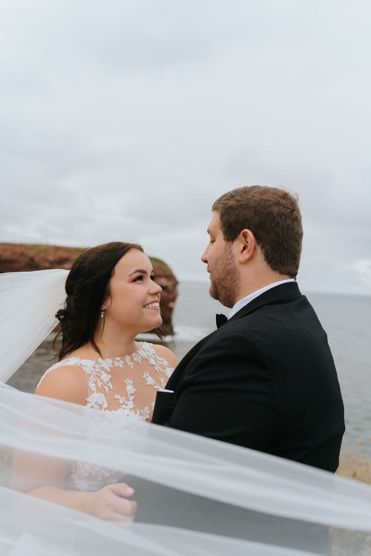 A couple in wedding attire share an intimate moment outdoors with a flowing veil in the foreground.