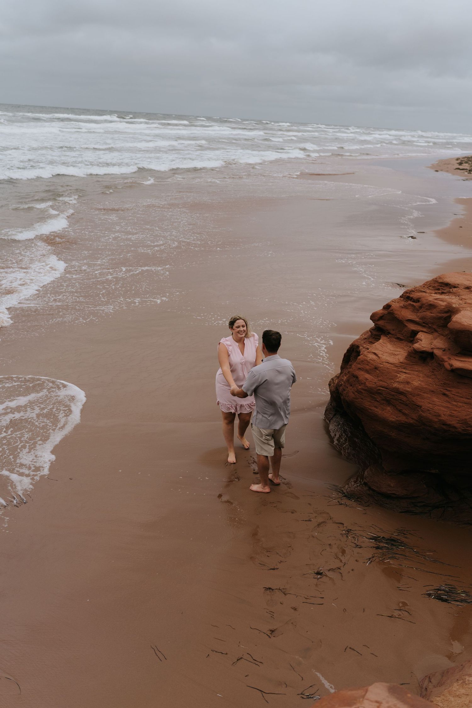 Two people explore red rock formations along a cloudy beach as waves crash against the shore at Thunder Cove PEI