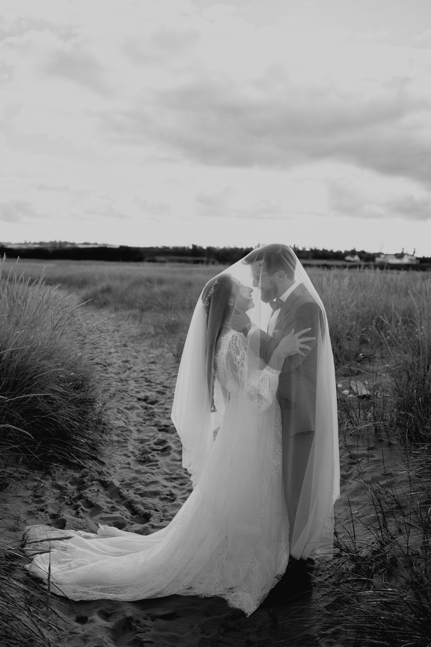 Dramatic black and white photo of couple sharing intimate moment under veil in coastal setting.