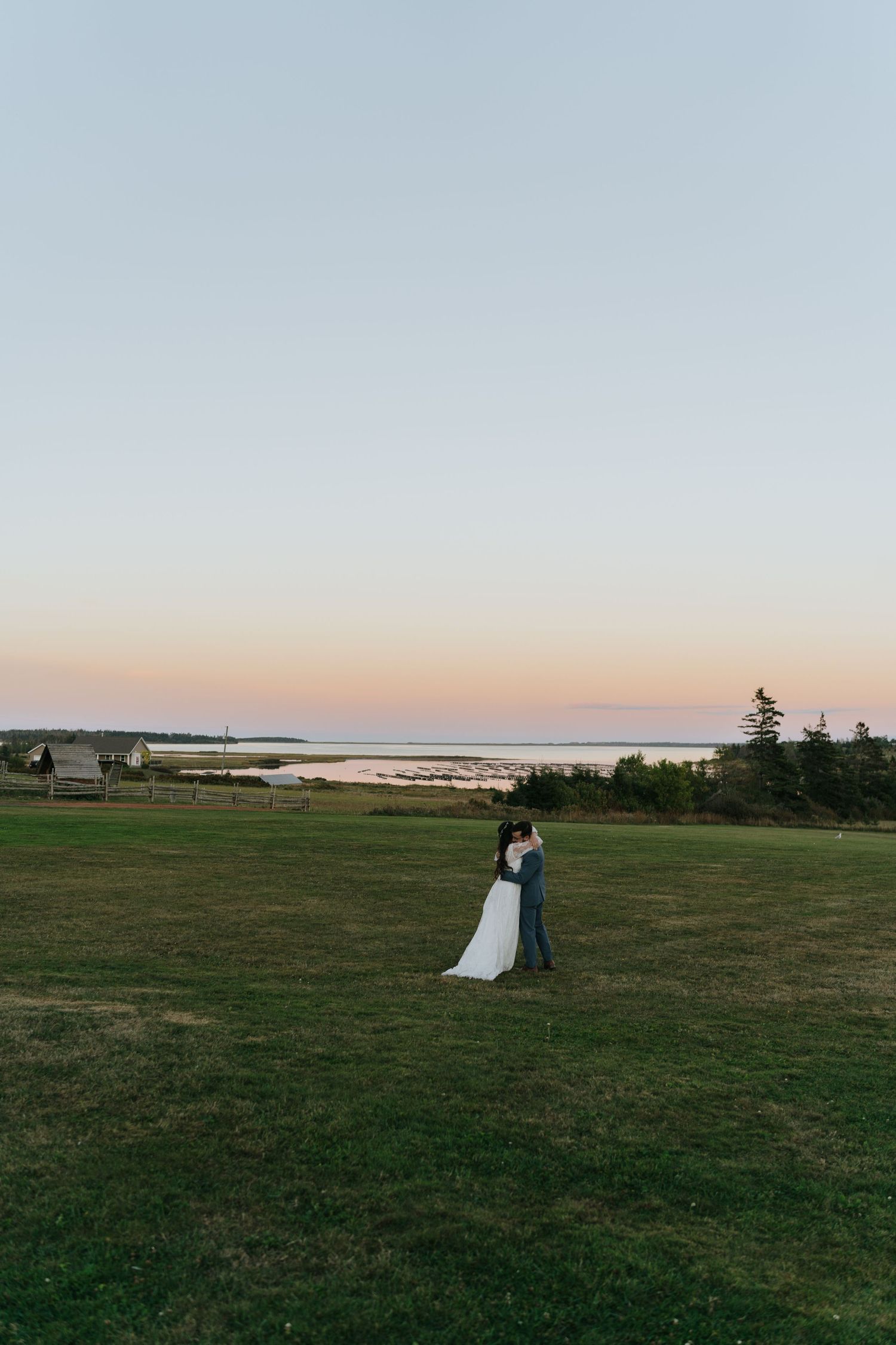 A couple in wedding attire stands in silhouette against a sunset on an open grassy field near the ocean.
