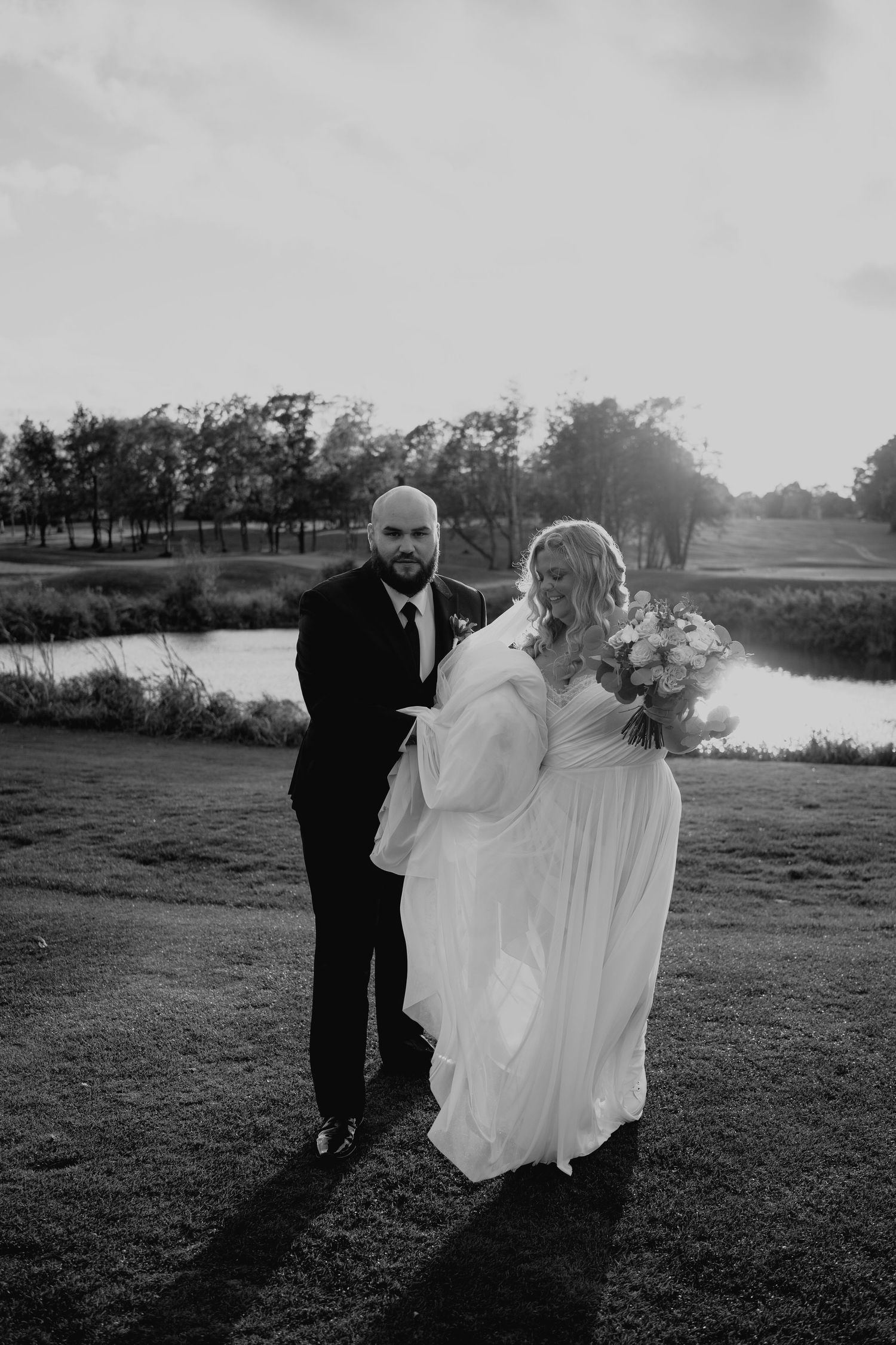 Black and white photo of newlyweds standing by a lake during golden hour with dramatic shadows.