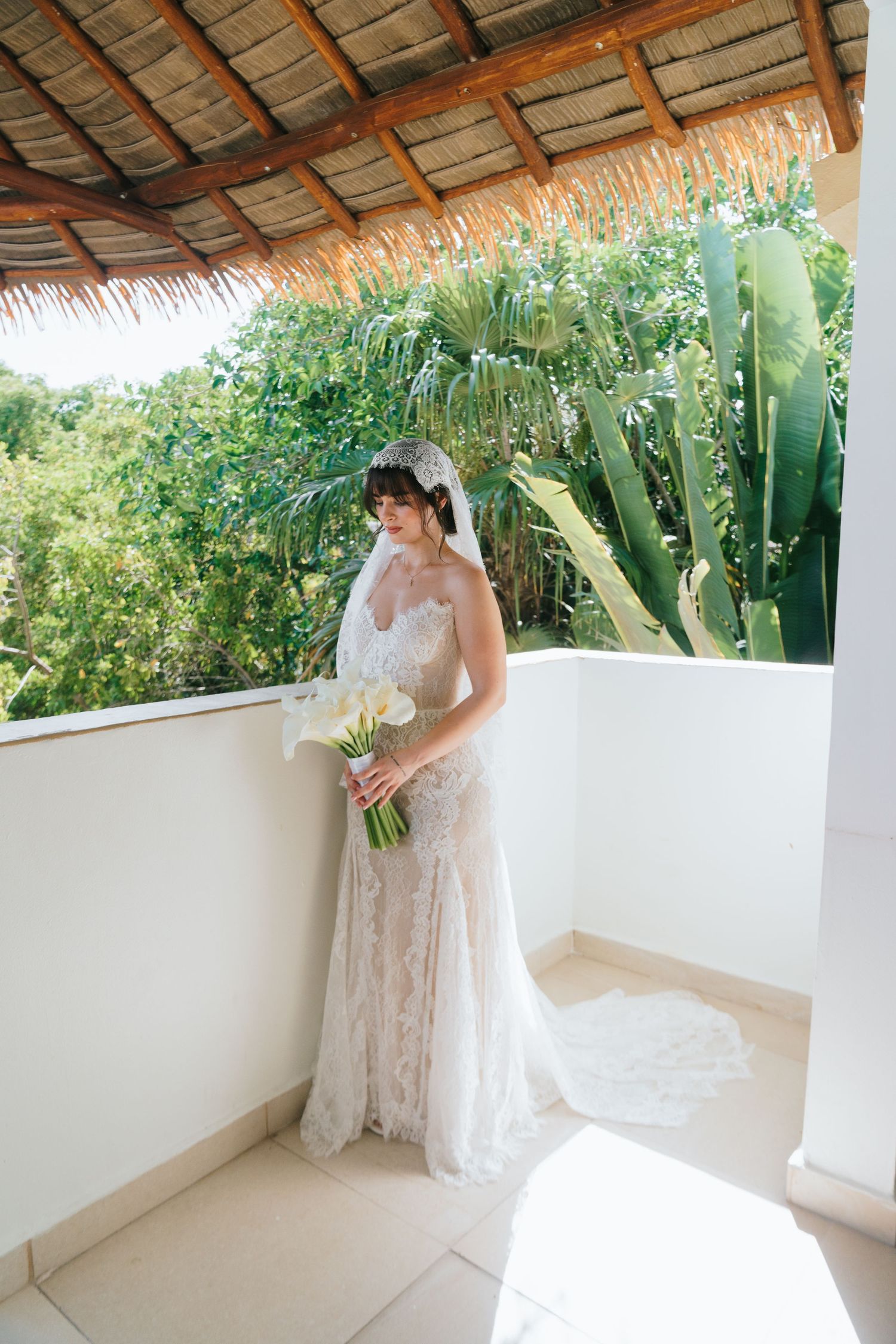Bride in lace gown and veil holding white calla lily bouquet on tropical balcony.
