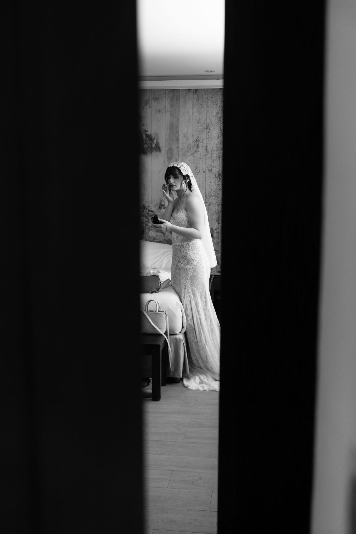 Bride in lace gown and veil seen through doorway in black and white photo.