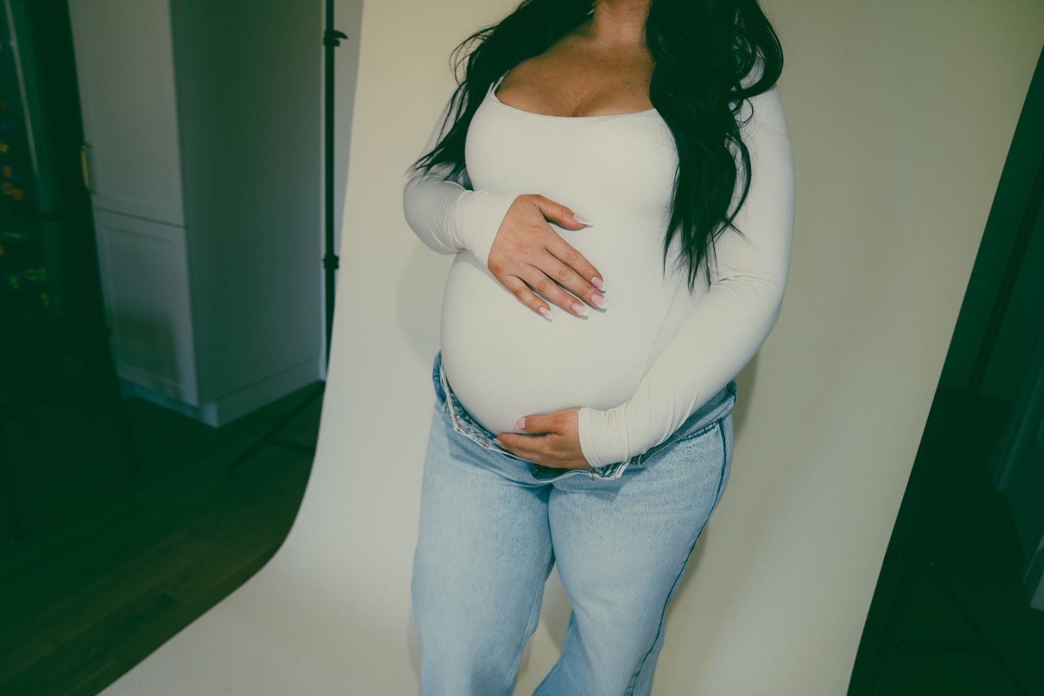 Pregnant woman in white top and jeans cradling baby bump during maternity photo shoot.