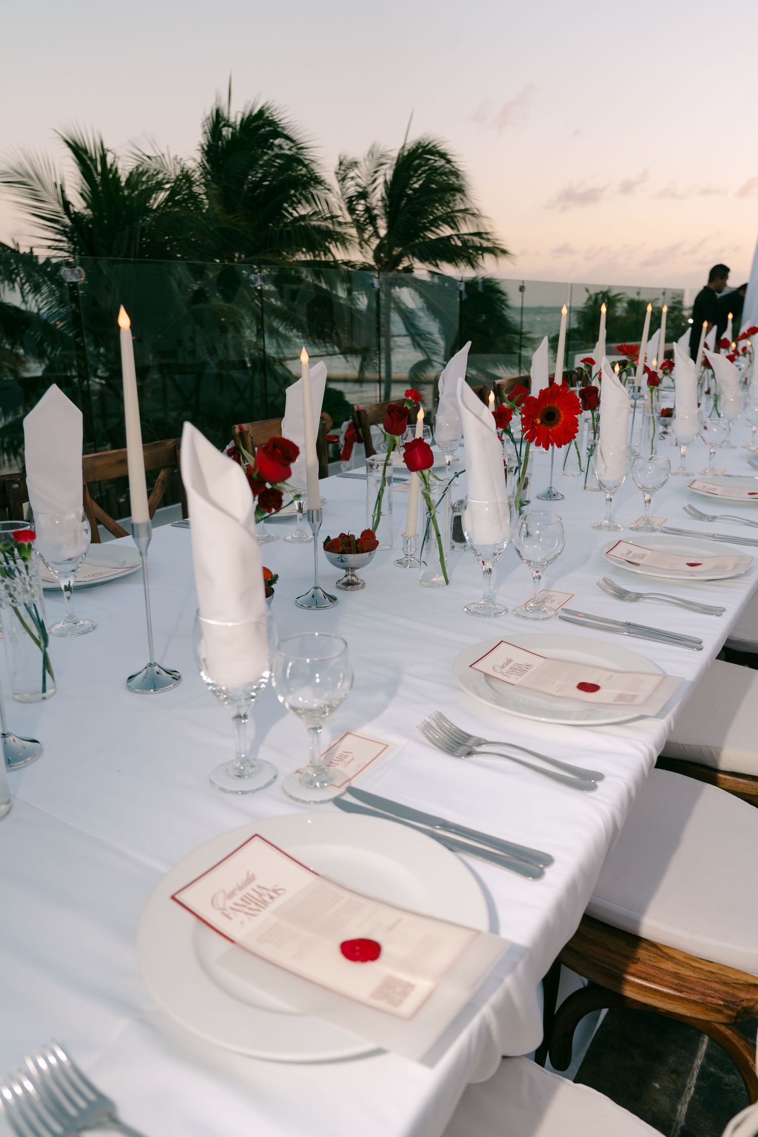 Formal wedding reception table set with white linens, red gerbera flowers, tall candles, and folded napkins outdoors.