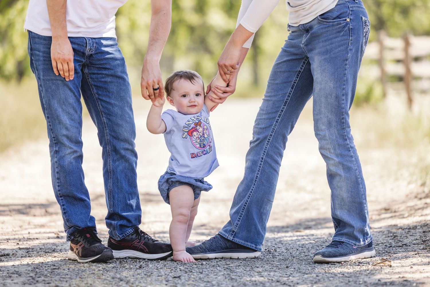 Two parents holding hands with a happy baby learning to walk on a gravel path outdoors.