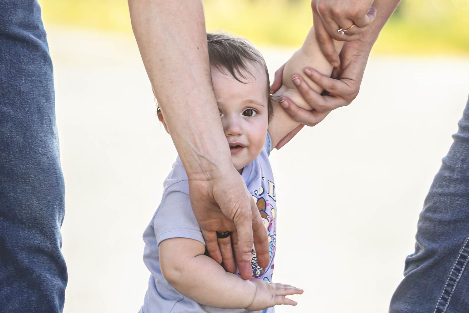 Baby learning to walk while being supported by two adults holding their hands outdoors.