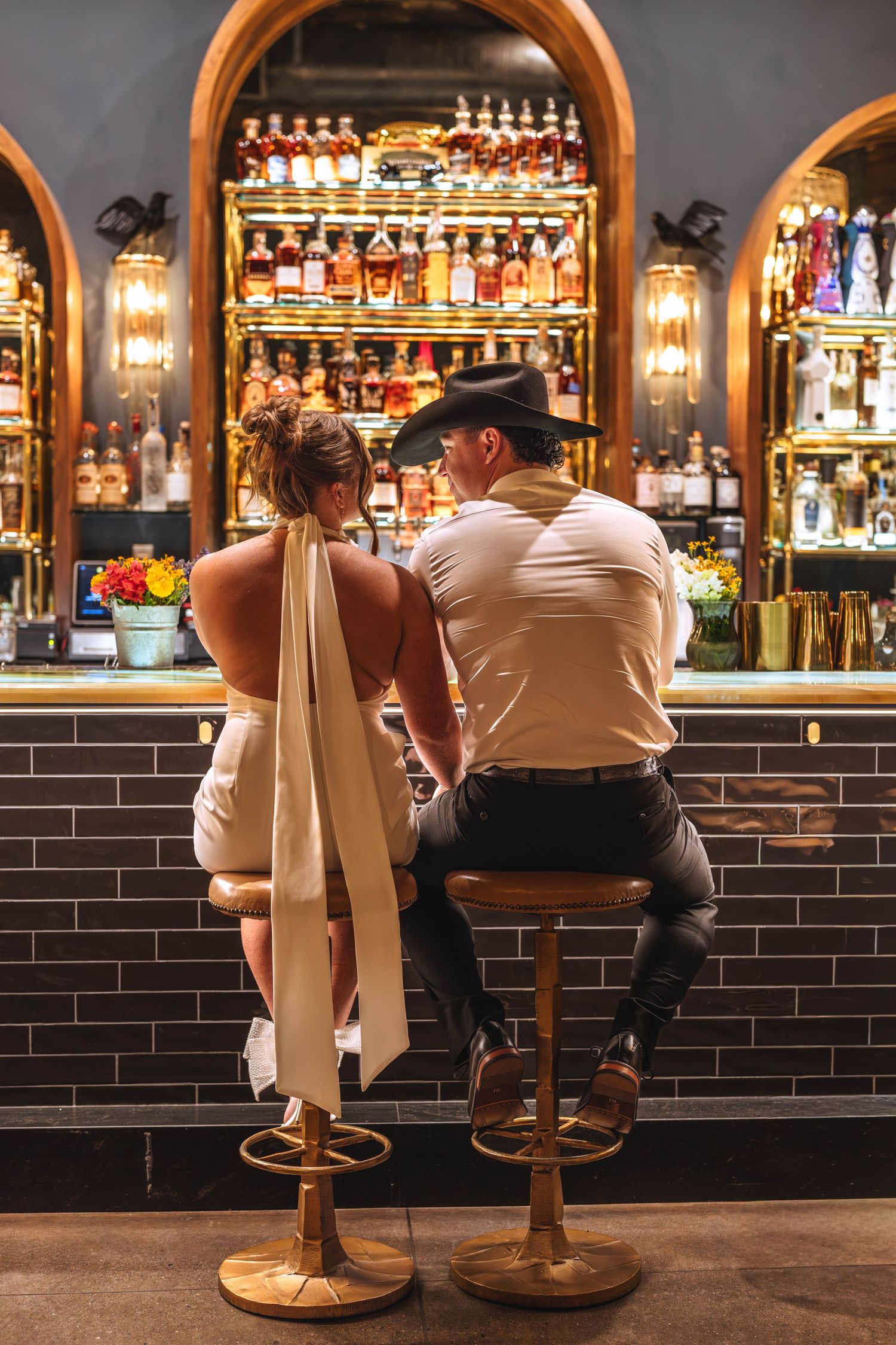 Couple in wedding attire sitting at a stylish bar with gold stools, surrounded by backlit shelves of spirits.