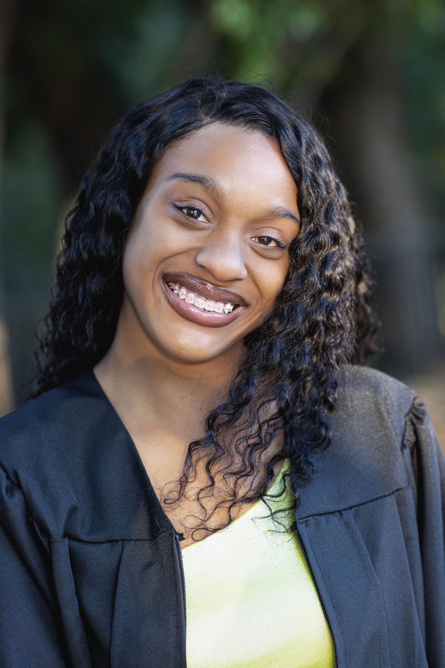 Professional portrait showing bright smile and curly hair against natural outdoor background.