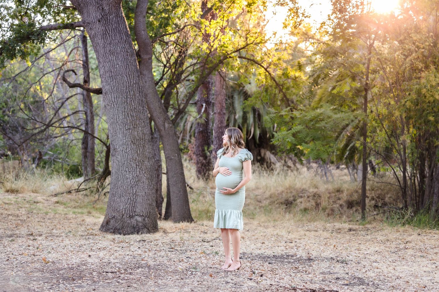 Pregnant woman in sage green dress standing outdoors among trees in golden hour light during maternity photo session.