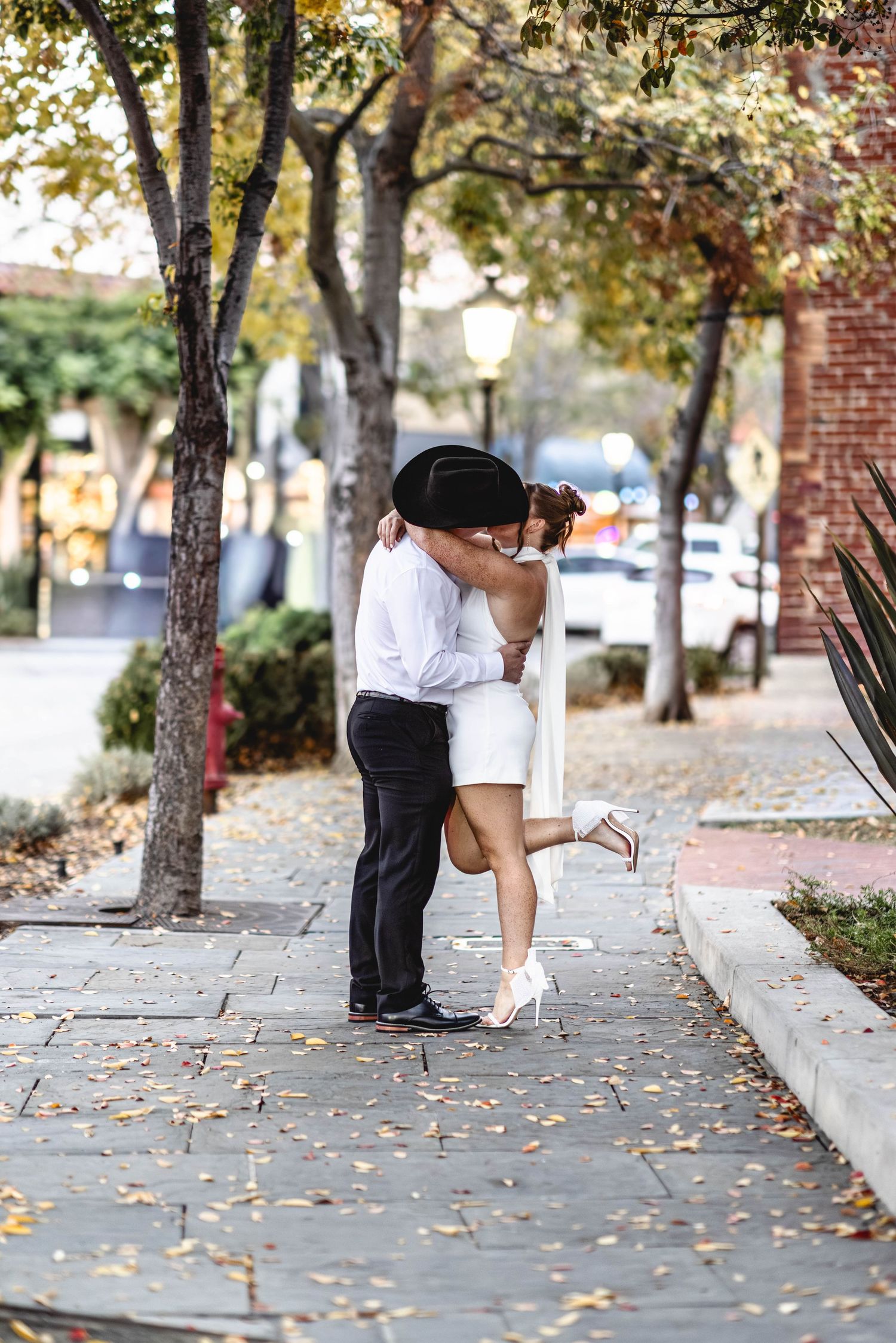 Couple sharing a romantic kiss on a tree-lined sidewalk in autumn, man in black hat and woman in white dress and heels.