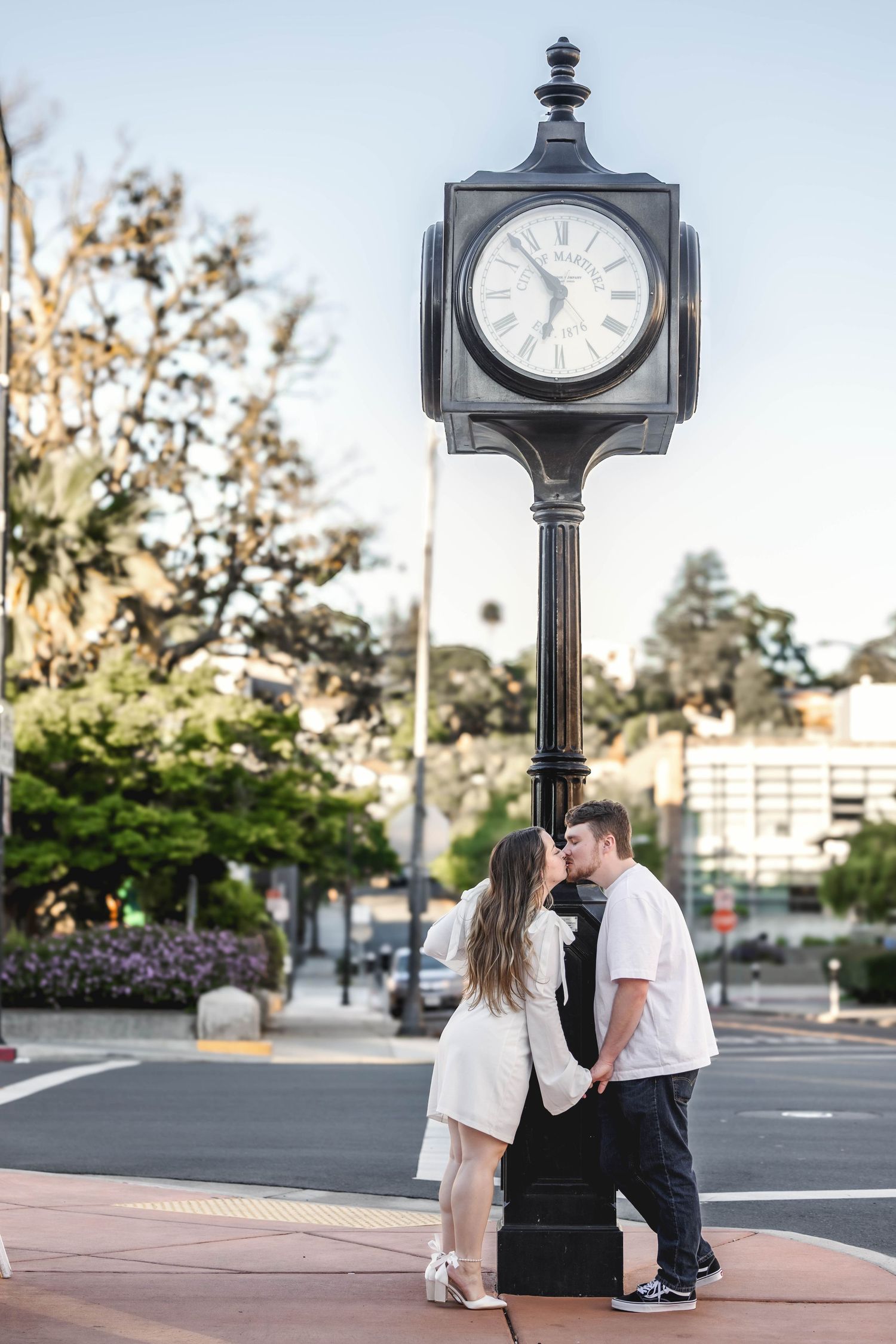 Couple sharing a kiss under a vintage street clock in a charming urban setting at golden hour.