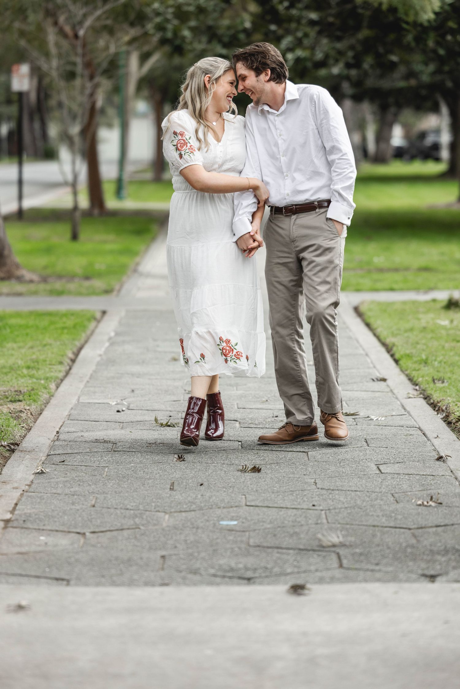 Couple sharing a romantic moment on a park pathway, woman in white floral dress with red boots, man in white shirt and khakis.