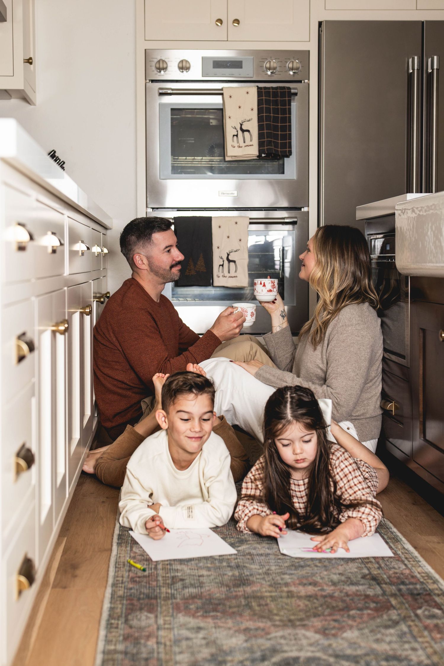 Family of four relaxing in a modern white kitchen, parents chatting over coffee while children draw on the floor.
