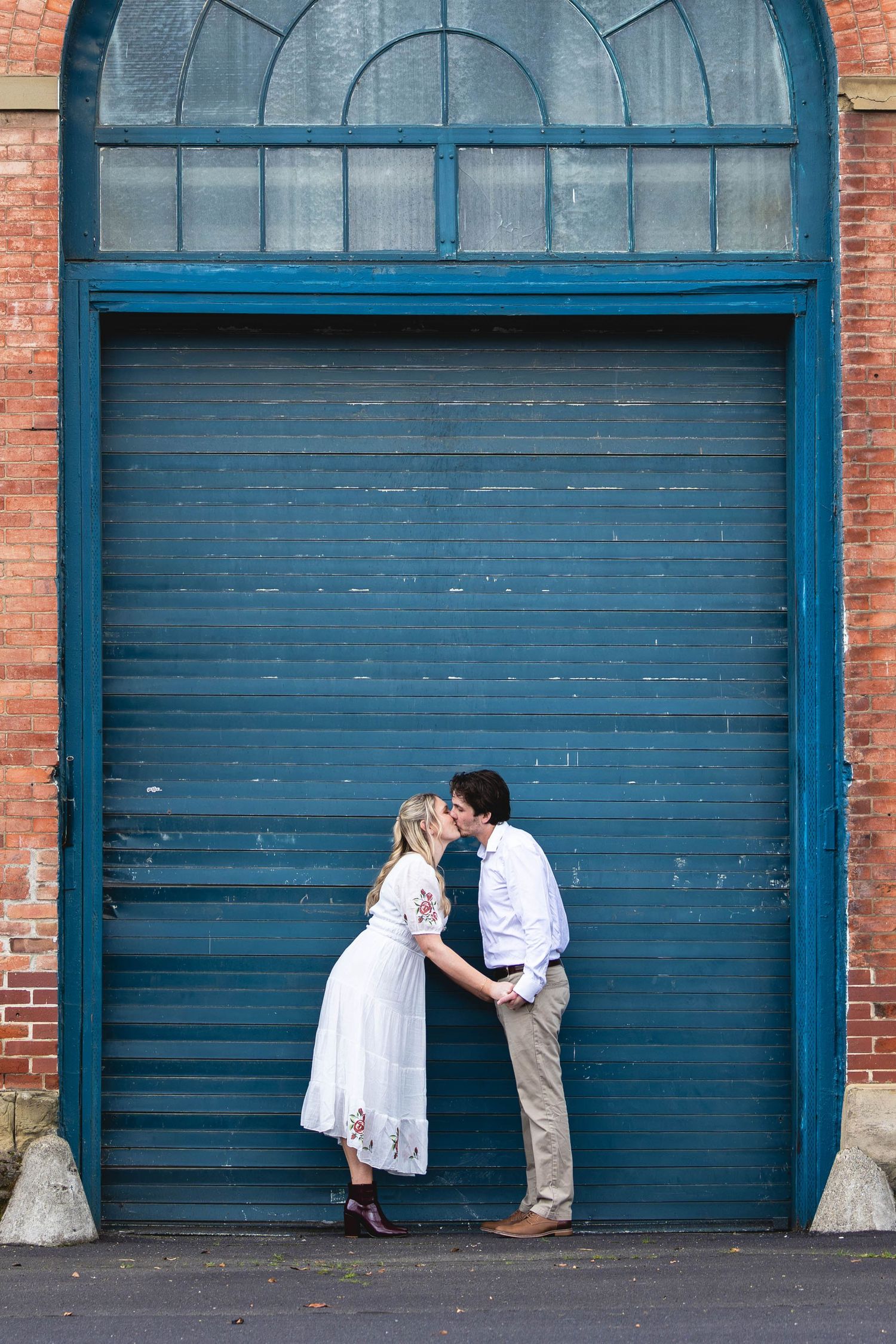 Couple sharing a romantic kiss in front of a blue wooden garage door with brick wall backdrop.