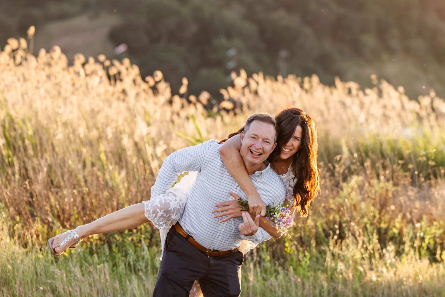 Couple laughing in golden field, man carrying woman on his back during sunny outdoor engagement photo session.