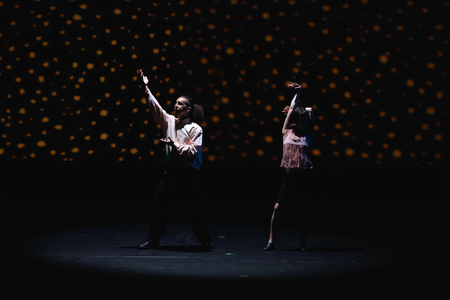 Two dancers perform on a dark stage with a golden polka dot backdrop, arms raised gracefully.