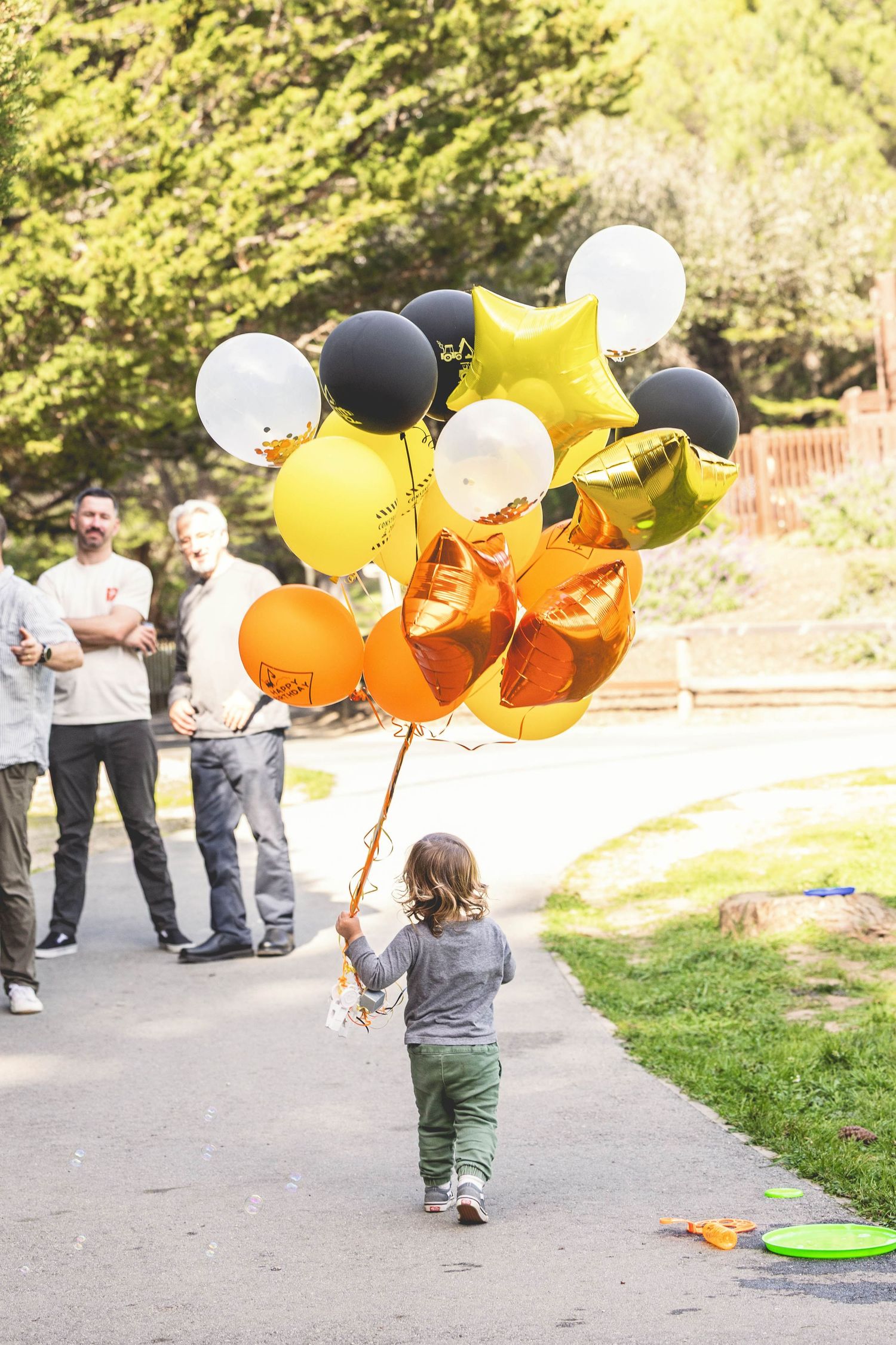 Young child walking away holding large bunch of black, gold, orange, and white balloons in sunny park.